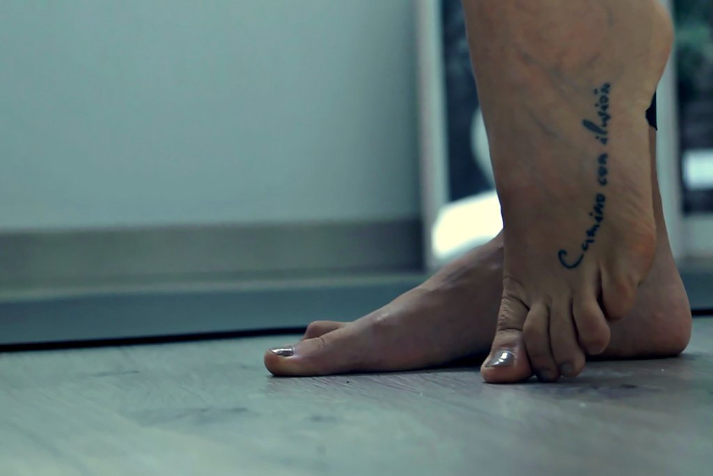 Dos pies descalzos apoyados en el suelo de madera del estudio, con una frase tatuada en el empeine