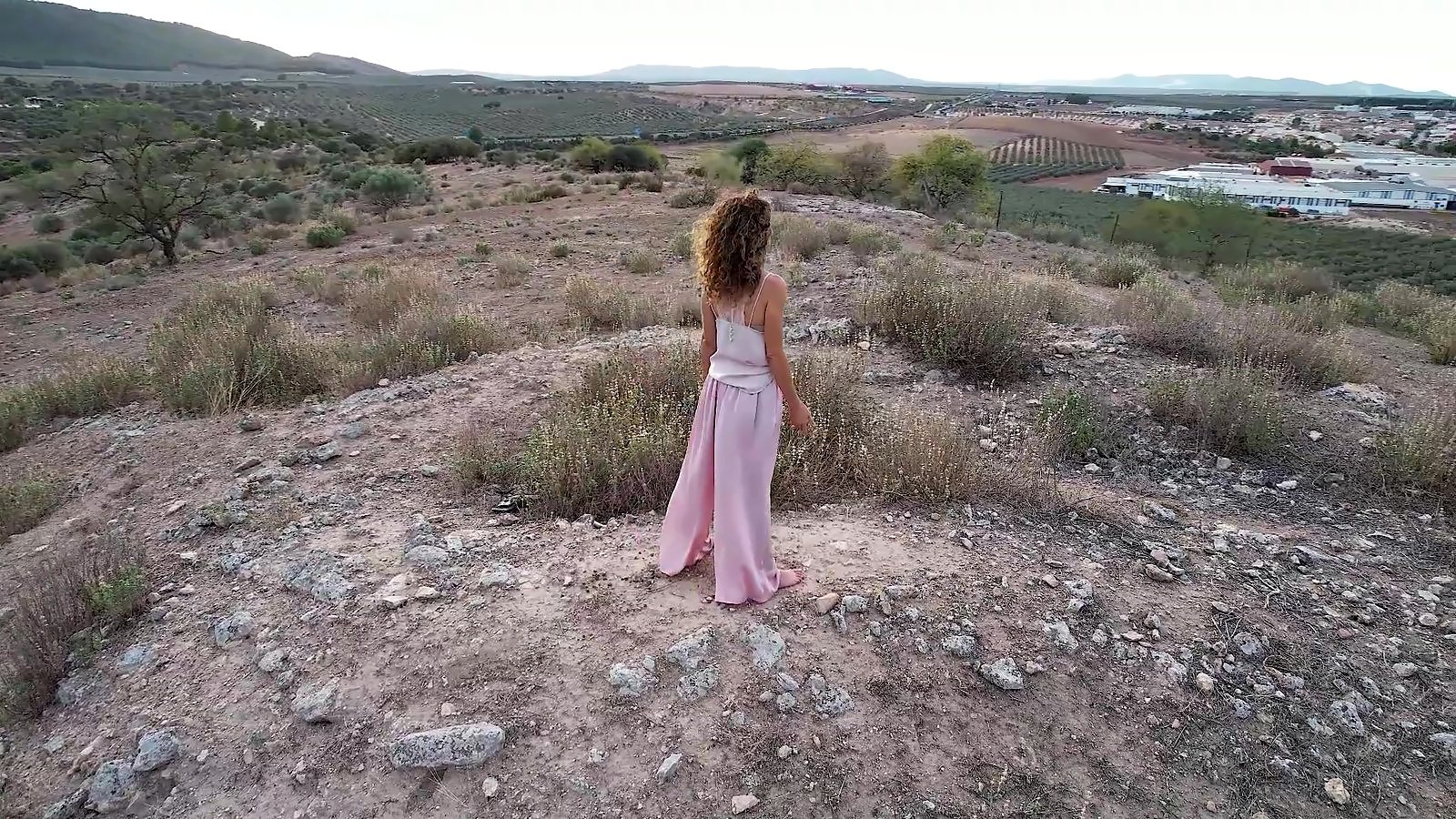 Ana, de espaldas, con pantalón rosa, en lo alto de un cerro; al fondo, Mollina, sus naves y los campos rojos