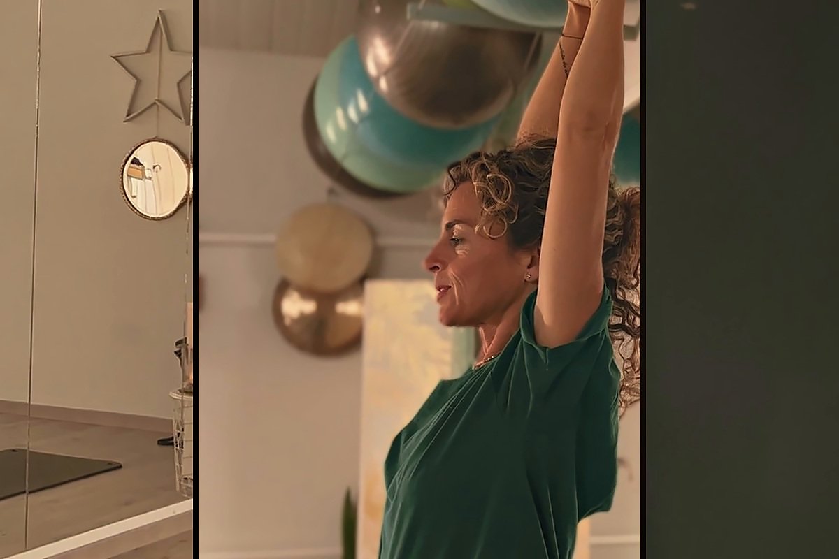 Ana, con la camiseta verde del estudio, estira los brazos hacia arriba en la sala, con pelotas grandes y dos gongs colgados detrás