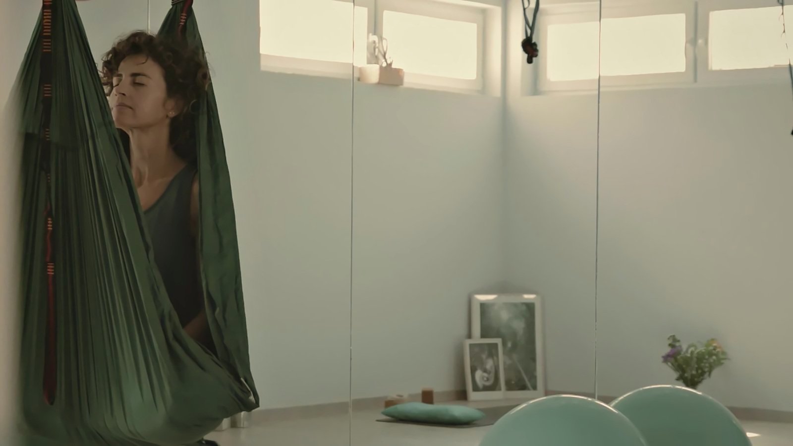 Ana, con los ojos cerrados, descansa suspendida en una hamaca de tela verde en la sala, con la luz de las ventanas altas